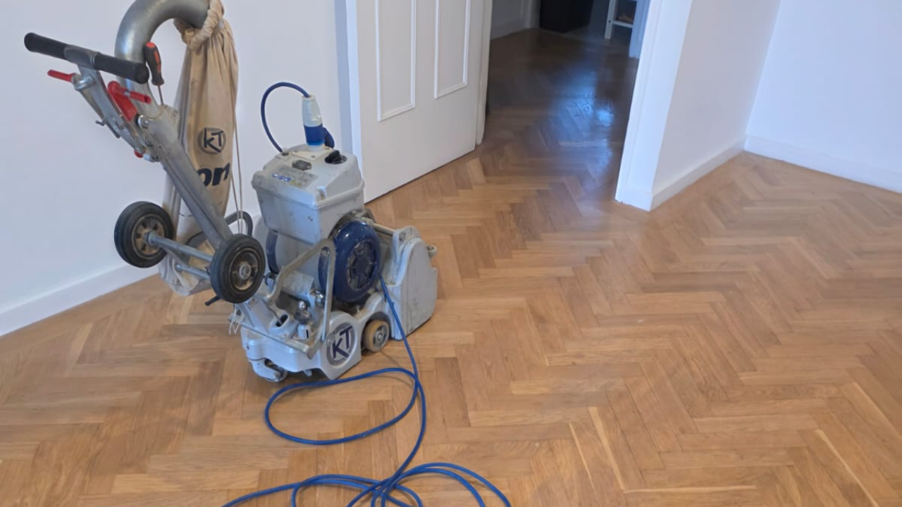 A contractor performing wood floor sanding on an oak parquet floor with a professional belt sander to reveal raw wood.