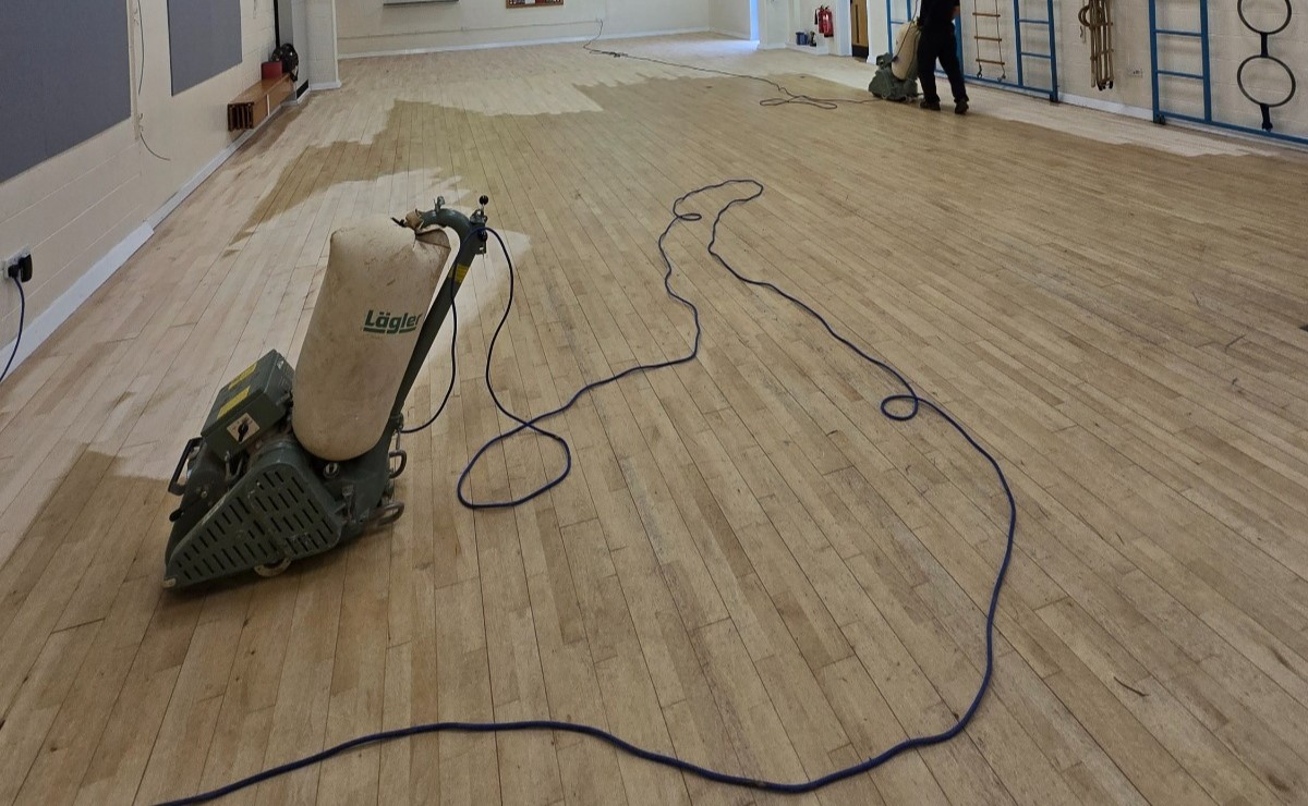 Commercial Floor Sanding in Carshalton on a sprung maple strip sports hall floor, showing belt sanders mid-way through restoring the worn surface.